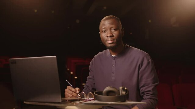 Medium portrait of young Black male comedian looking at camera while sitting by laptop at dark amateur theater writing jokes for his standup concert