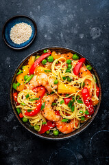 Stir fry noodles with shrimps, colorful paprika, green pea, chives and sesame seeds with ginger, garlic and soy sauce. Black table background, top view