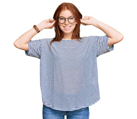 Young read head woman wearing casual clothes and glasses smiling pulling ears with fingers, funny...