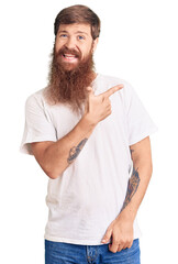 Handsome young red head man with long beard wearing casual white tshirt cheerful with a smile of face pointing with hand and finger up to the side with happy and natural expression on face