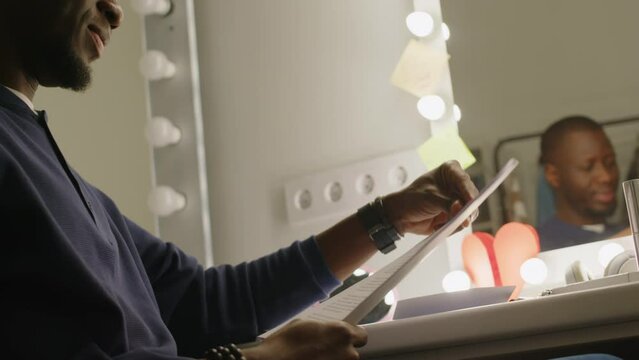 Low angle of Black man reading his monologue and laughing at jokes while getting ready for standup concert in backstage room with light bulb mirror