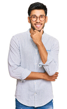 Young handsome man wearing casual clothes and glasses looking confident at the camera smiling with crossed arms and hand raised on chin. thinking positive.