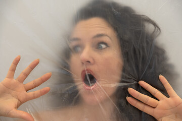 blurred portrait of a brunette curly woman behind transparent plastic foil with defensive hands, mouth opened to scream, as a symbol of fear, panic, worry and despair