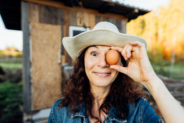 Laughing Woman in Hat Holds Egg to Face, Closing Eye . Fresh chicken egg in the female hands of a farmer, rural life, organic farming Happy moment of collecting eggs