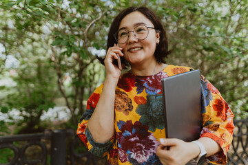 Smiling freelancer woman in a laptop using a smartphone outdoors in the park. Brightly dressed woman with technical gadgets calling, talking on a mobile phone surrounded by green trees in the park