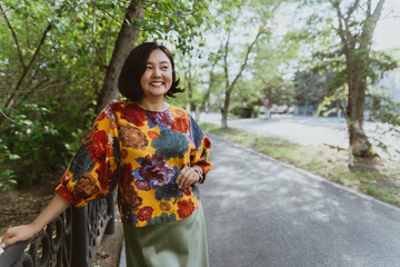 Stylish lady wearing a trendy, floral patterned top stands by park railing Vibrant portrait of a cheerful Asian woman with a great smile outdoors