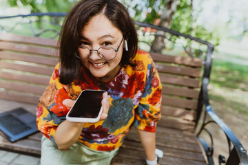 Smiling freelancer woman wearing colorful blouse, animatedly chatting on her smartphone outdoor. Entrepreneur with digital devices, passionately engaged in loud phone discussion in urban green space