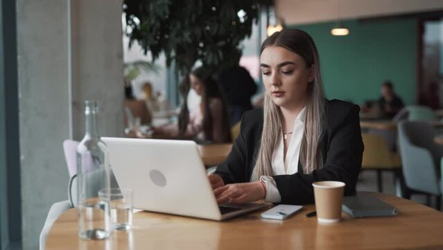 beautiful, stylish young woman sits in a cafe in business attire, opens her laptop, and starts working with a smile
