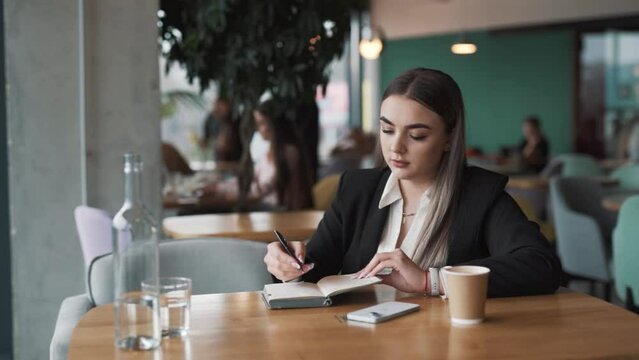 stylish young woman sits in a cafe in business attire, writing in a notebook with a pen, smiling