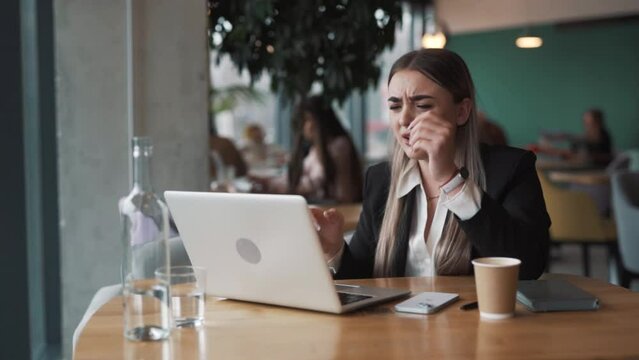 beautiful, sad young woman sits in a cafe in business attire, saddened by what she sees on her laptop, grasping her head
