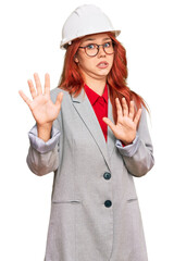 Young redhead woman wearing architect hardhat afraid and terrified with fear expression stop gesture with hands, shouting in shock. panic concept.