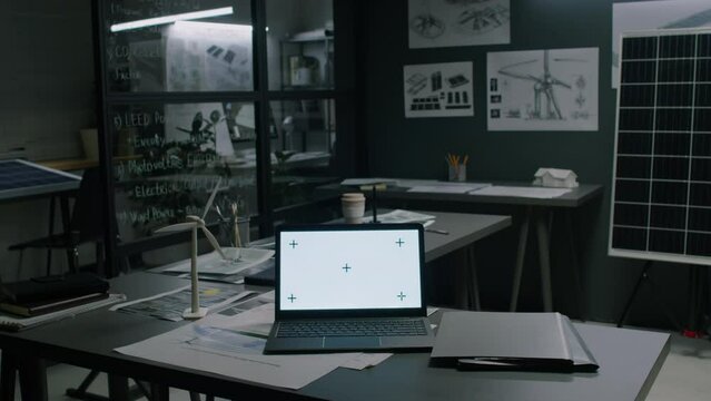 No people footage of interior of empty alternative energy department office with multiple blueprints and drawings, laptop with chromakey screen on table, windmill and solar panel models