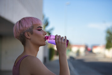 Portrait of young, attractive, gay, heavily makeup man with pink hair and top, drinking water from a bottle on a hot afternoon. LGTBIQ+ concept, gay, pride, makeup, fashion, trend.