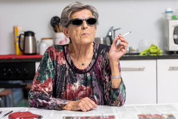 Portrait of old smoker sitting in her modern white kitchen with dark sunglasses. A woman from another time, A beautiful lady full of wrinkles.