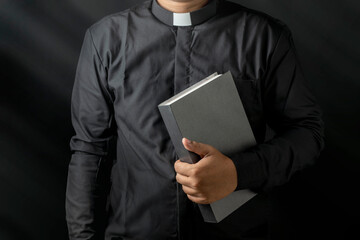 Young priest holding bible isolated on black background. - obrazy, fototapety, plakaty
