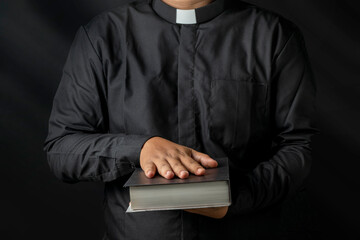 Young priest holding bible and other hand covering  isolated on black background. - obrazy, fototapety, plakaty