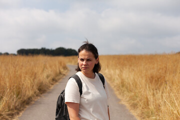 Young woman with backpack standing on the road in the field, looking at camera