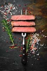 Poster Slices of beef steak on a metal fork on a dark background. Top view. Free space for text. © Yaruniv-Studio