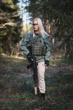 Beautiful military blonde girl with a modern AK 12 suppressed rifle in the forest. Vertical photo.