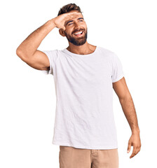 Young hispanic man wearing casual white tshirt very happy and smiling looking far away with hand over head. searching concept.