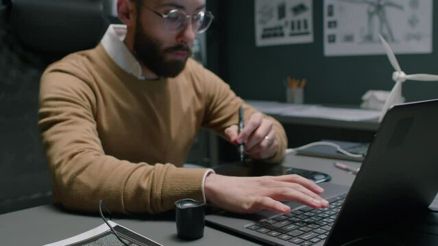 Tilt footage of male Middle Eastern engineer typing data into laptop and drawing on graphic tablet while working on project in renewable energy department office at night