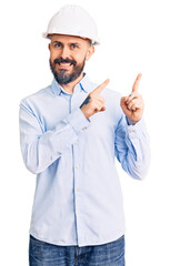 Young handsome man wearing architect hardhat smiling and looking at the camera pointing with two hands and fingers to the side.