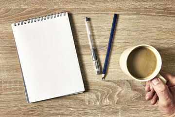 an open notebook with a pencil and a hand holding a cup of coffee on a wooden table, top view