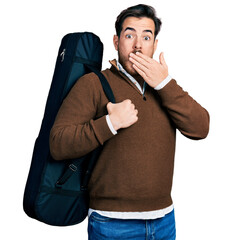 Young hispanic man wearing guitar case covering mouth with hand, shocked and afraid for mistake....
