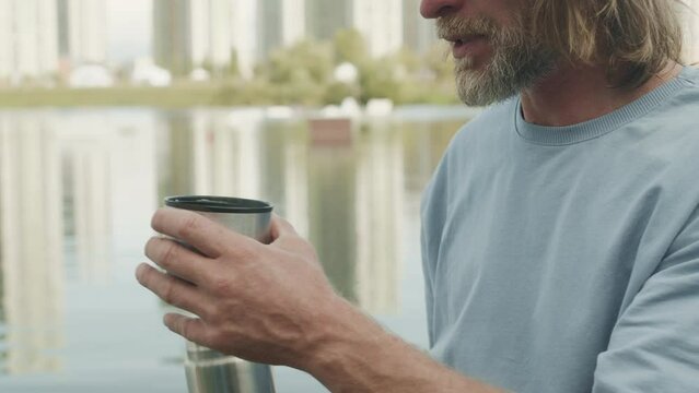 Chest up slowmo of Caucasian adult man pouring and drinking hot tea from thermos while sitting by water in peaceful city park with view