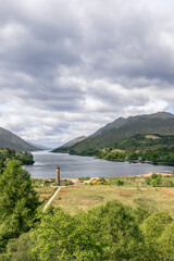 Vertically framed, the Glenfinnan Monument rises beside the serene Loch Shiel, drawing the eye along the water to the distant Scottish Highlands under a broad sky