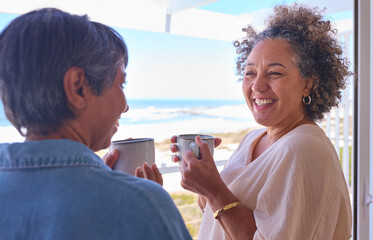 Two Mature Women Friends In Beachfront House Overlooking Ocean For Summer Vacation With Hot Drinks
