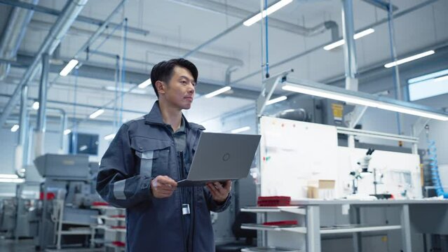 Chief Engineer Walking Through the Hall in a Modern Factory with Manufacturing Machinery and Production Tools. Asian Specialist Holding a Laptop Computer. Successful Man in an Industrial Environment