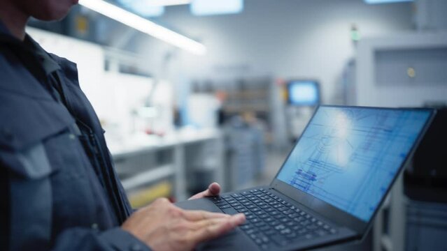 Electronics Factory Worker Analyzing Detailed Engineering Blueprints on a Laptop Computer Screen. Over the Shoulder Arc Footage with Adult Asian Man Working a Modern Industrial Job