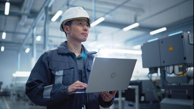 Portrait of an Asian Industrial Specialist Wearing Uniform and Protective Hard Hat in Electronics Manufacturing Facility. Professional Technician Using Laptop Computer while Walking in the Hallway