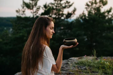 Foto op Canvas Young woman sitting in lotus position with tibetan singing bowl meditating on rocky mountain at sunset. © dsheremeta