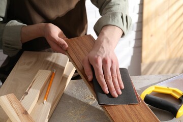 Man polishing wooden plank with sandpaper at grey table indoors, closeup - obrazy, fototapety, plakaty