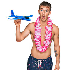 Young blond man wearing swimsuit and hawaiian lei holding airplane toy scared and amazed with open...