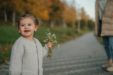 Baby girl holding a bouquet of wild flowers picked from the park, smiling as her mother waits for her. Happy family walk.Joyful little girl