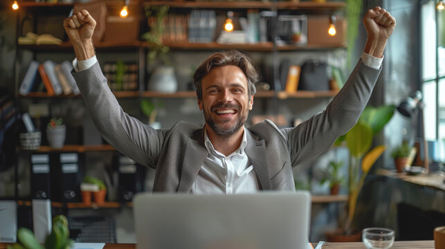 A man sits in front of a laptop with his hands raised in joy.