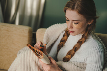 Young minded woman wrapped in plaid writing down memories in notebook sitting on sofa at home...