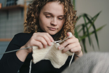 Young curly haired woman knitting handmade piece while sitting on couch at home and enjoying cozy hobby. Handmade clothes. The girl goes in for her hobbies