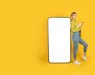 Using mobile application, young full body length blonde girl leaning big huge blank screen phone with white touchscreen mock up. Using smartphone, looking camera. Yellow background, copy space.