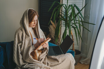 Image of a young woman wrapped in cozy blanket taking notes on her notebook and using her laptop computer to manage her tasks at home.