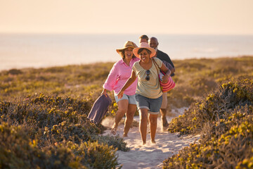 Mature Couple With Friends On Summer Vacation Walking Through Dunes On Way To Beach Carrying Bags