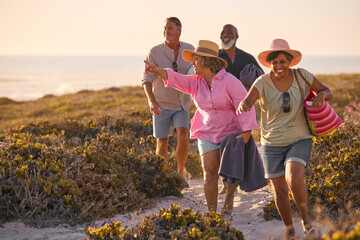Mature Couple With Friends On Summer Vacation Walking Through Dunes On Way To Beach Carrying Bags