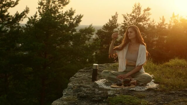 A young woman meditating with a smoking palo santo in her hands while sitting on a rock at sunset.