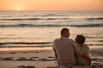 Rear View Of Mature Couple Sitting On Beach Shoreline Looking Out To Sea At Sunset