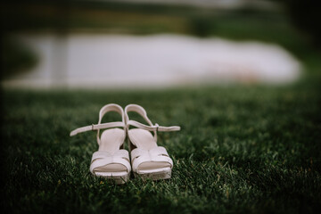 Elegant white bridal shoes on a grassy field with a blurred water body in the background, possibly...