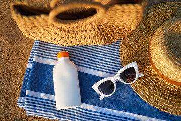 Sunscreen bottle on striped towel next to woven bag, straw hat, and white sunglasses on sandy beach...