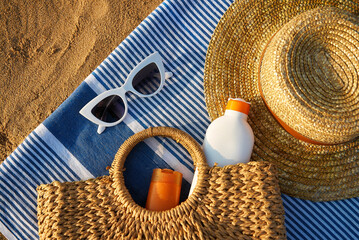 Summer accessories on sandy beach at sunset. Woven bag with sunscreen inside next to stylish sun hat. White sunglasses lie on blue striped towel. Vacation essentials for sun protection.